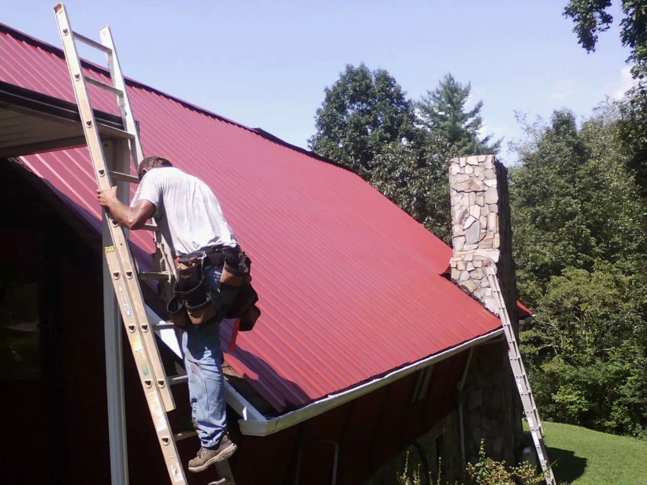 Roofer on a ladder beside a red metal roof and stone chimney in White Haven
